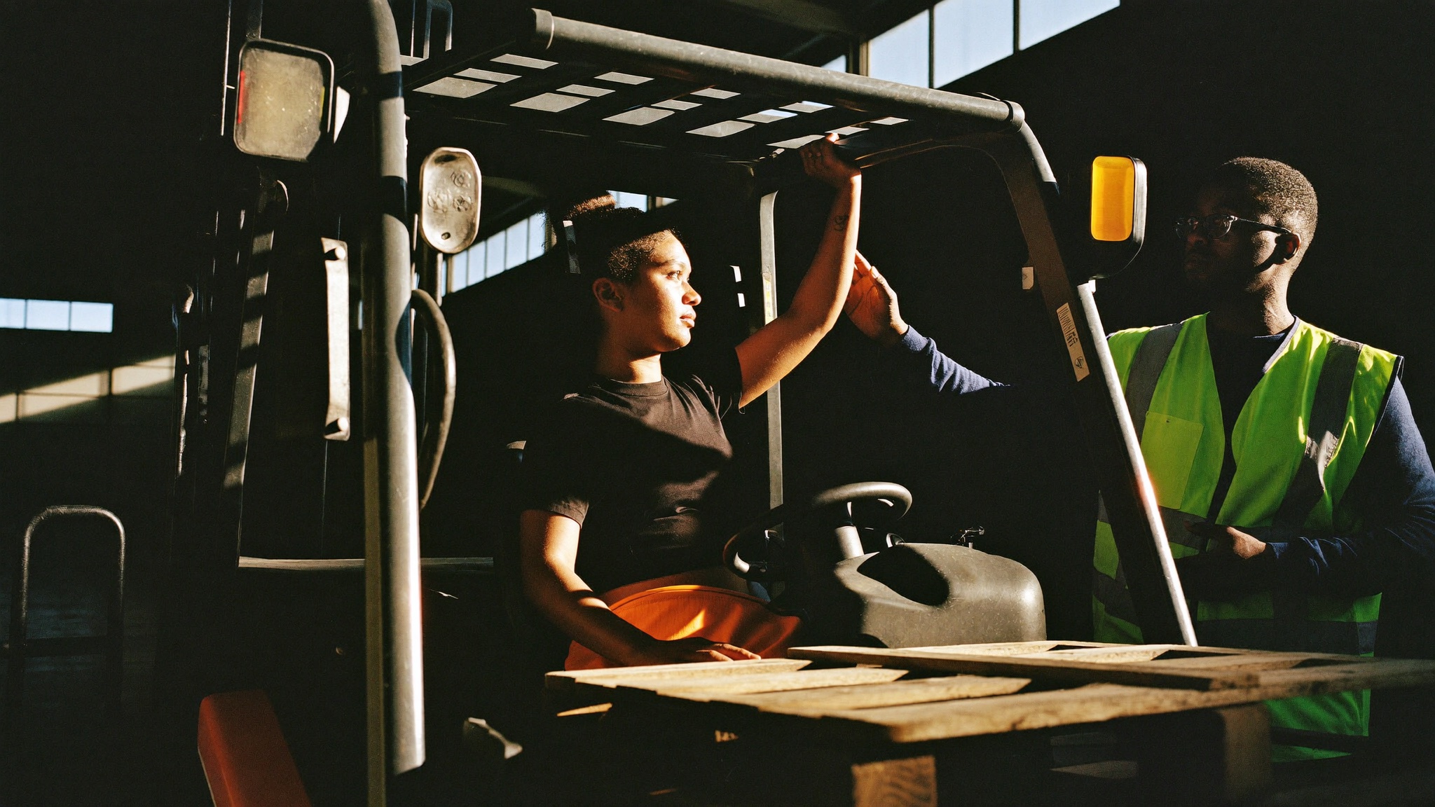 A young woman learning forklift operation in a warehouse training bay, an instructor guiding her in golden light